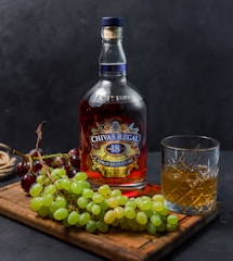 A bottle of Chivas Regal 18 Gold Signature whiskey is placed on a wooden board next to a glass filled with whiskey and ice. Bunches of green and red grapes accompany the arrangement, adding a touch of freshness. The background is a dark, textured surface that creates a contrast with the vibrant colors of the objects in the foreground.