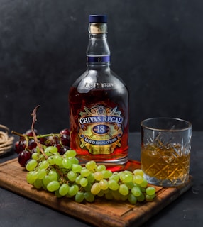 A bottle of Chivas Regal 18 Gold Signature whiskey is placed on a wooden board next to a glass filled with whiskey and ice. Bunches of green and red grapes accompany the arrangement, adding a touch of freshness. The background is a dark, textured surface that creates a contrast with the vibrant colors of the objects in the foreground.