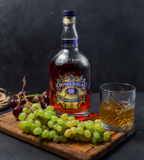 A bottle of Chivas Regal 18 Gold Signature whiskey is placed on a wooden board next to a glass filled with whiskey and ice. Bunches of green and red grapes accompany the arrangement, adding a touch of freshness. The background is a dark, textured surface that creates a contrast with the vibrant colors of the objects in the foreground.