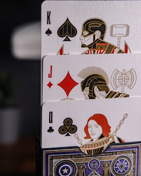 Three playing cards featuring intricate designs and illustrations of figures resembling superheroes. The cards displayed are the King of Spades, the Jack of Diamonds, and the Queen of Clubs. Each card shows a uniquely illustrated character with detailed symbolic armor and weapons.