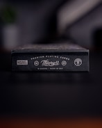 A close-up view of a black box of premium playing cards with the brand names Marvel and Theory11 prominently displayed. The design includes sleek typography and logo symbols, conveying a sense of luxury and exclusivity. The box is situated on a dark surface with a blurred background.