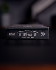 A close-up view of a black box of premium playing cards with the brand names Marvel and Theory11 prominently displayed. The design includes sleek typography and logo symbols, conveying a sense of luxury and exclusivity. The box is situated on a dark surface with a blurred background.