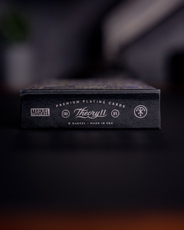 A close-up view of a black box of premium playing cards with the brand names Marvel and Theory11 prominently displayed. The design includes sleek typography and logo symbols, conveying a sense of luxury and exclusivity. The box is situated on a dark surface with a blurred background.