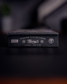 A close-up view of a black box of premium playing cards with the brand names Marvel and Theory11 prominently displayed. The design includes sleek typography and logo symbols, conveying a sense of luxury and exclusivity. The box is situated on a dark surface with a blurred background.