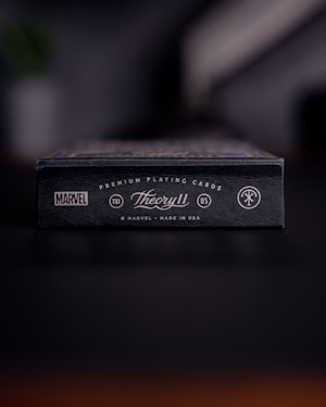 A close-up view of a black box of premium playing cards with the brand names Marvel and Theory11 prominently displayed. The design includes sleek typography and logo symbols, conveying a sense of luxury and exclusivity. The box is situated on a dark surface with a blurred background.