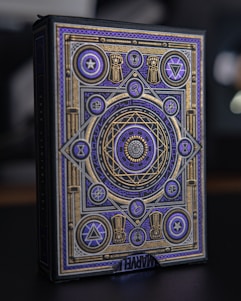 An ornate box with intricate geometric and symbolic patterns in gold and purple adorns its surface. Various symbols and shapes such as stars, triangles, and circles are incorporated into the design. The patterns are symmetric and appear to convey a mystical or magical theme. The word 'MARVEL' is partially visible at the bottom.