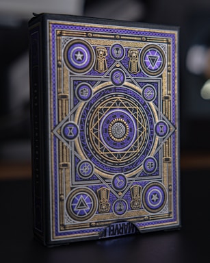 An ornate box with intricate geometric and symbolic patterns in gold and purple adorns its surface. Various symbols and shapes such as stars, triangles, and circles are incorporated into the design. The patterns are symmetric and appear to convey a mystical or magical theme. The word 'MARVEL' is partially visible at the bottom.