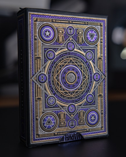 An ornate box with intricate geometric and symbolic patterns in gold and purple adorns its surface. Various symbols and shapes such as stars, triangles, and circles are incorporated into the design. The patterns are symmetric and appear to convey a mystical or magical theme. The word 'MARVEL' is partially visible at the bottom.