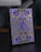 A box of playing cards adorned with intricate designs and symbols. The central focus is a large 'A' surrounded by circular emblems and geometric patterns. The design includes a combination of gold and violet hues, with various symbols often associated with superhero themes.