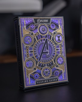 A box of playing cards adorned with intricate designs and symbols. The central focus is a large 'A' surrounded by circular emblems and geometric patterns. The design includes a combination of gold and violet hues, with various symbols often associated with superhero themes.