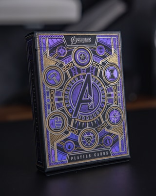 A box of playing cards adorned with intricate designs and symbols. The central focus is a large 'A' surrounded by circular emblems and geometric patterns. The design includes a combination of gold and violet hues, with various symbols often associated with superhero themes.
