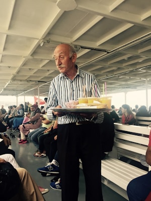 An elderly man with a striped shirt holds a tray with drinks and snacks in a busy indoor seating area. He stands near rows of benches filled with people, some attentively watching him while others are engaged in conversations.