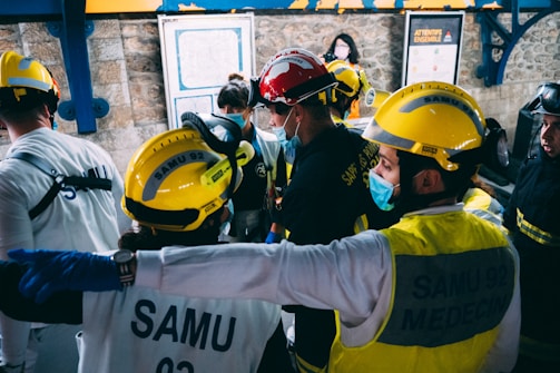 A group of emergency medical personnel wearing helmets and protective gear are gathered in what looks like a train station or industrial area. They appear to be engaged in discussion or planning, with one individual pointing forward. The background features stone walls, a map, and informational signs.