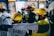 A group of emergency medical personnel wearing helmets and protective gear are gathered in what looks like a train station or industrial area. They appear to be engaged in discussion or planning, with one individual pointing forward. The background features stone walls, a map, and informational signs.