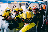 A group of miners in training wearing helmets and safety gear, attentively listening to an instructor outdoors.
