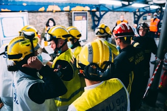 A group of rescue workers equipped with yellow and red helmets are gathered indoors. Some are wearing masks and uniforms with visible text, possibly indicating a coordinated operation or training session. The setting appears to be a structured, possibly industrial environment with other individuals in the background.
