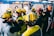 A group of rescue workers equipped with yellow and red helmets are gathered indoors. Some are wearing masks and uniforms with visible text, possibly indicating a coordinated operation or training session. The setting appears to be a structured, possibly industrial environment with other individuals in the background.