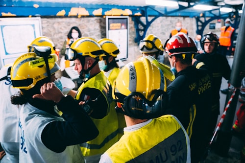 A group of rescue workers equipped with yellow and red helmets are gathered indoors. Some are wearing masks and uniforms with visible text, possibly indicating a coordinated operation or training session. The setting appears to be a structured, possibly industrial environment with other individuals in the background.