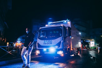 A truck with emergency lights is parked on a dimly lit street at night. A person wearing a face mask and dark clothing walks in front of the truck. The truck is marked with the words 'SAMU IVECO' on its front. Surrounding the scene, there are street signs and a few buildings with lights illuminating parts of the area.