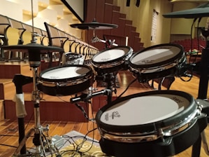 A set of electronic drums is positioned in an auditorium with tiered seating in the background. The room is spacious with wooden flooring and a series of black and white chairs organized in rows. The drum set, equipped with cymbals and multiple drum pads, is centrally placed on what appears to be a stage area.