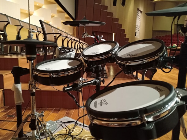 A set of electronic drums is positioned in an auditorium with tiered seating in the background. The room is spacious with wooden flooring and a series of black and white chairs organized in rows. The drum set, equipped with cymbals and multiple drum pads, is centrally placed on what appears to be a stage area.