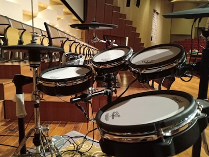 A set of electronic drums is positioned in an auditorium with tiered seating in the background. The room is spacious with wooden flooring and a series of black and white chairs organized in rows. The drum set, equipped with cymbals and multiple drum pads, is centrally placed on what appears to be a stage area.