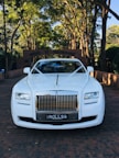 A luxurious white car is adorned with elegant wedding decorations, parked on a brick driveway under a canopy of tall trees. The background features a brick archway leading to a lush garden.