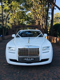 A luxurious white car is adorned with elegant wedding decorations, parked on a brick driveway under a canopy of tall trees. The background features a brick archway leading to a lush garden.