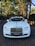 A luxurious white car is adorned with elegant wedding decorations, parked on a brick driveway under a canopy of tall trees. The background features a brick archway leading to a lush garden.