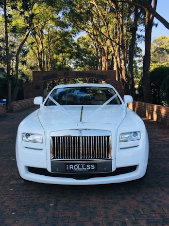 A luxurious white car is adorned with elegant wedding decorations, parked on a brick driveway under a canopy of tall trees. The background features a brick archway leading to a lush garden.