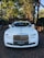 A luxurious white car is adorned with elegant wedding decorations, parked on a brick driveway under a canopy of tall trees. The background features a brick archway leading to a lush garden.