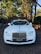 A luxurious white car is adorned with elegant wedding decorations, parked on a brick driveway under a canopy of tall trees. The background features a brick archway leading to a lush garden.