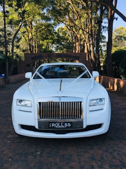 A luxurious white car is adorned with elegant wedding decorations, parked on a brick driveway under a canopy of tall trees. The background features a brick archway leading to a lush garden.