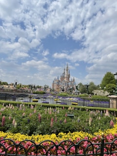 A beautiful fairy-tale castle is set in the background under a sky with scattered clouds. The foreground features a vibrant garden filled with an array of colorful flowers, creating a picturesque and enchanting scene.