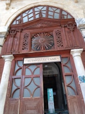 An intricately carved wooden entrance with ornate geometric and floral patterns, featuring a semicircular arch with frosted glass panels above. A sign with the name 'Kütahya Ulu Camii' is displayed over the doorway, which is framed by stone columns.
