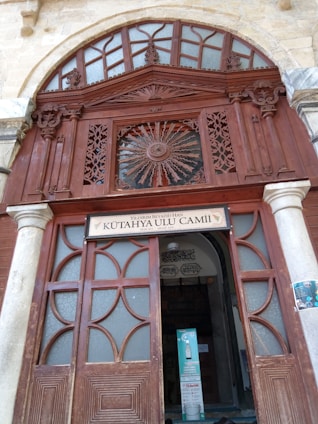 An intricately carved wooden entrance with ornate geometric and floral patterns, featuring a semicircular arch with frosted glass panels above. A sign with the name 'Kütahya Ulu Camii' is displayed over the doorway, which is framed by stone columns.