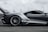 A sleek, high-performance sports car moving at speed. The car has a modern aerodynamic design with a prominent rear spoiler and a predominantly metallic color scheme. The blurred background suggests motion, highlighting the car's dynamic presence on what appears to be a racetrack.