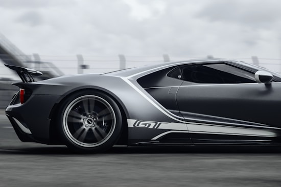A sleek, high-performance sports car moving at speed. The car has a modern aerodynamic design with a prominent rear spoiler and a predominantly metallic color scheme. The blurred background suggests motion, highlighting the car's dynamic presence on what appears to be a racetrack.