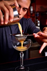 A bartender carefully pours a yellow cocktail mixture through a fine mesh strainer into a coupe glass. The focus is on the hands and the cocktail, capturing the precision and skill involved in the preparation. The bartender is in a dimly lit bar setting.