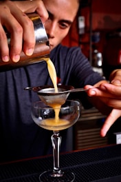 A bartender carefully pours a yellow cocktail mixture through a fine mesh strainer into a coupe glass. The focus is on the hands and the cocktail, capturing the precision and skill involved in the preparation. The bartender is in a dimly lit bar setting.
