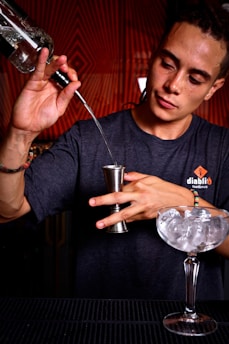A person is concentrating while pouring a liquid from a bottle into a jigger. Ice-filled cocktail glass is placed on a bar counter. The person is wearing a dark t-shirt and has a focused expression. The background features a patterned red design, creating a warm atmosphere.