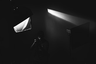 A black and white image depicting a person holding a camera in what appears to be a dimly lit studio setup. Softbox lighting equipment is present, casting a beam of light across the scene, and creating dramatic shadows.