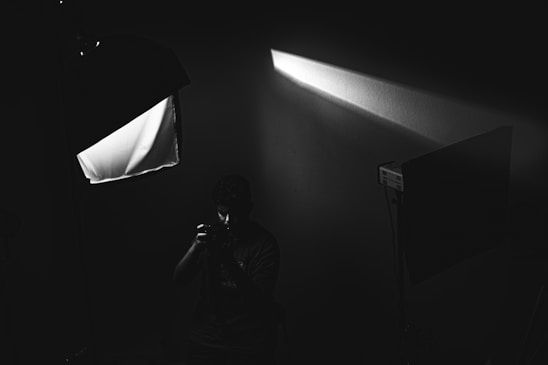 A black and white image depicting a person holding a camera in what appears to be a dimly lit studio setup. Softbox lighting equipment is present, casting a beam of light across the scene, and creating dramatic shadows.