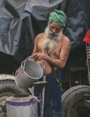 An elderly man with a long white beard and a green turban is pouring liquid from a metal bucket into a container through a funnel. He is shirtless, wearing a dark blue cloth around his waist. The background features large, dark tarpaulin and vehicle wheels.