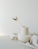 A minimalist ceramic vase sits atop a soft, light-colored fabric with a single dried grass stem extending from it. The background is a smooth, neutral shade, creating a clean and calming composition.