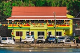A building with a sign reading 'GTA Grenada Tourism Authority' is seen with a colorful facade featuring green, red, and yellow stripes. The building has a red roof, and a string of multicolored triangular flags is strung in front. In front of the building, several parked vehicles, including cars and a truck, are visible. A few people are walking or standing near the vehicles. The background includes greenery with bright flowers on a hillside.