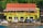 A building with a sign reading 'GTA Grenada Tourism Authority' is seen with a colorful facade featuring green, red, and yellow stripes. The building has a red roof, and a string of multicolored triangular flags is strung in front. In front of the building, several parked vehicles, including cars and a truck, are visible. A few people are walking or standing near the vehicles. The background includes greenery with bright flowers on a hillside.