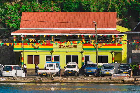 A building with a sign reading 'GTA Grenada Tourism Authority' is seen with a colorful facade featuring green, red, and yellow stripes. The building has a red roof, and a string of multicolored triangular flags is strung in front. In front of the building, several parked vehicles, including cars and a truck, are visible. A few people are walking or standing near the vehicles. The background includes greenery with bright flowers on a hillside.