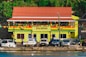 A building with a sign reading 'GTA Grenada Tourism Authority' is seen with a colorful facade featuring green, red, and yellow stripes. The building has a red roof, and a string of multicolored triangular flags is strung in front. In front of the building, several parked vehicles, including cars and a truck, are visible. A few people are walking or standing near the vehicles. The background includes greenery with bright flowers on a hillside.