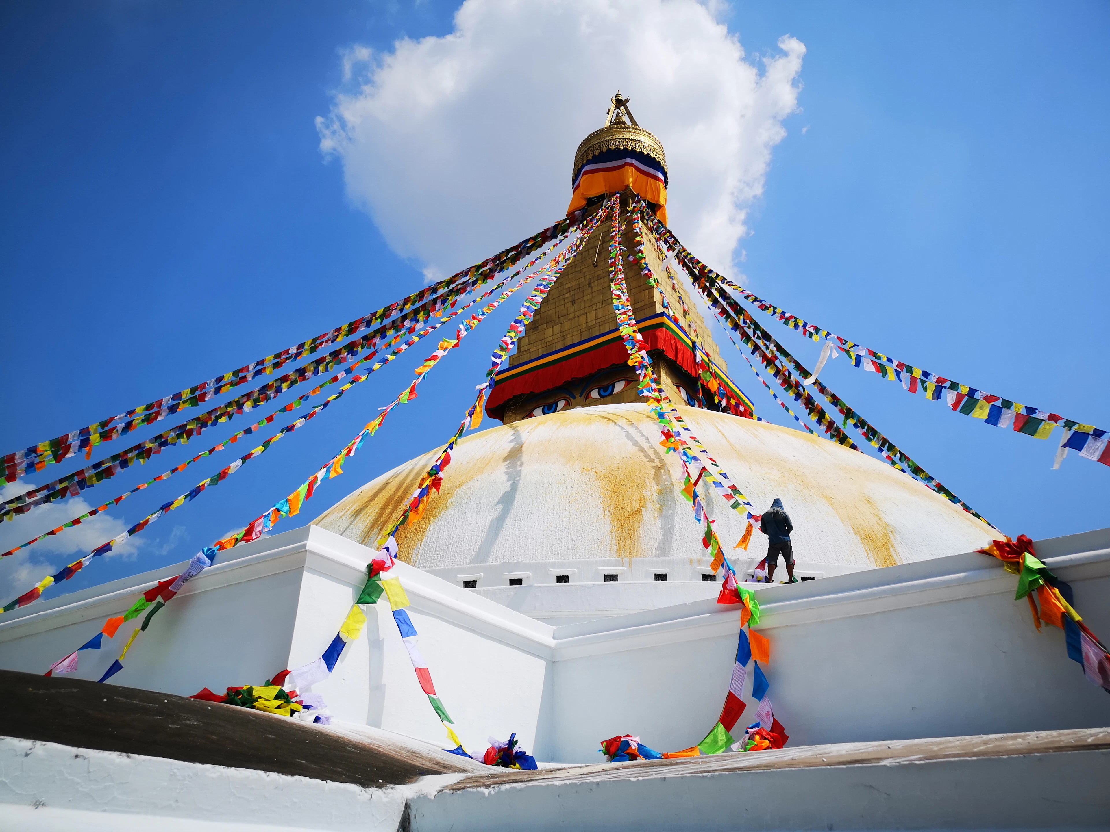 A lone figure walks along Boudhanath Stupa's base as prayer flags radiate from its golden spire under a cloudy sky.
